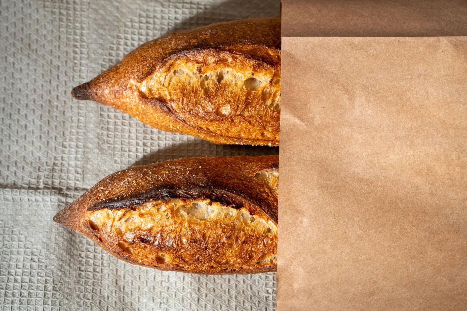 Two dark-baked baguettes half wrapped in a plain brown paper bag
