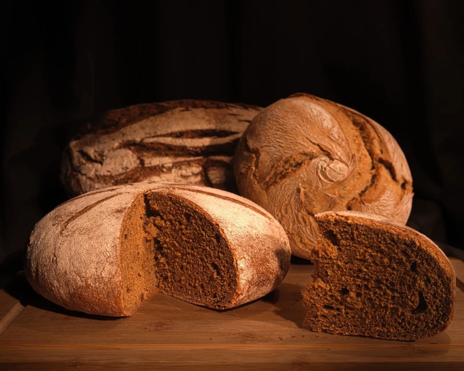 Dark rye loaves on a wooden board, one cut open to show a dense brown crumb