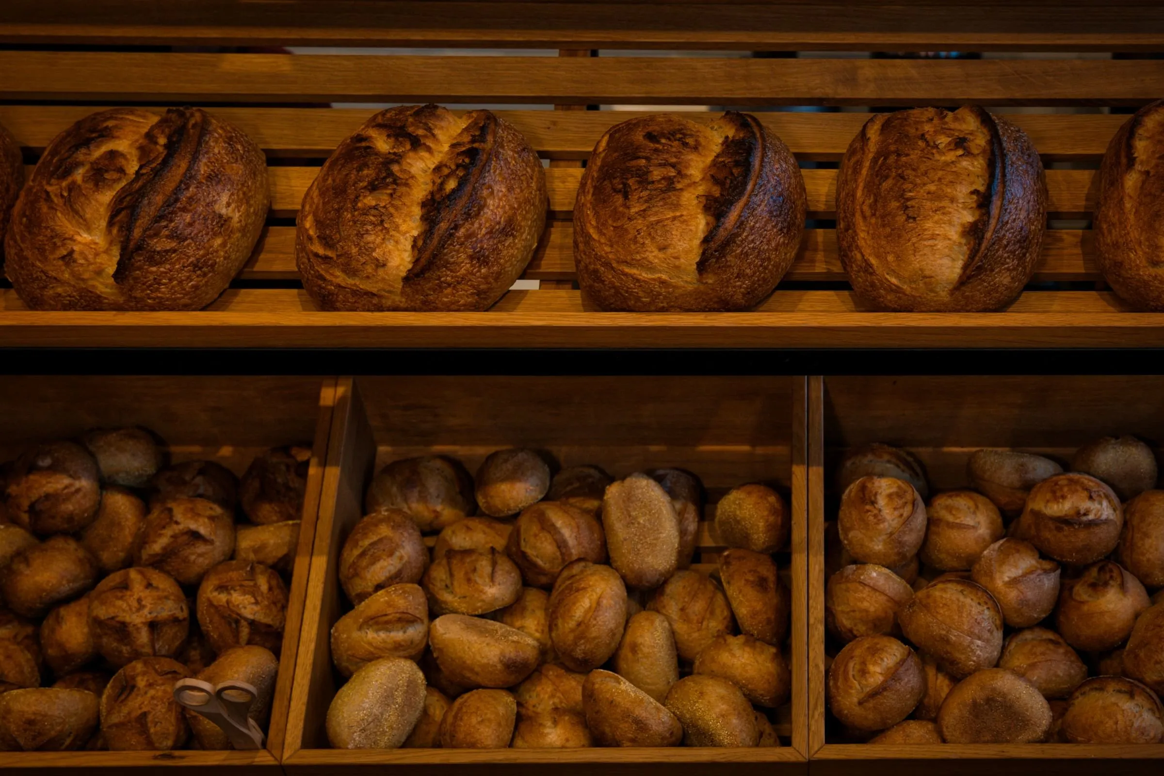 Scored sourdough loaves on an oak shelf above wooden crates of crusty rolls